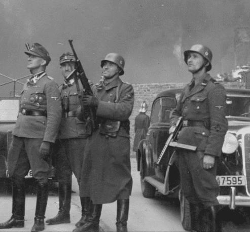 Stroop Report original caption- "The leader of the grand operation." SS-Brigadeführer Jürgen Stroop (center) watches housing blocks burn. The SD-Rottenführer at right is Josef Blösche ("Frankenstein"). Photo taken at Nowolipie street looking east, near the intersection with Smocza street. On the left is the burning balcony of the townhouse at Nowolipie 66; next to it is the Ghetto wall. (courtesy wikipedia.org)