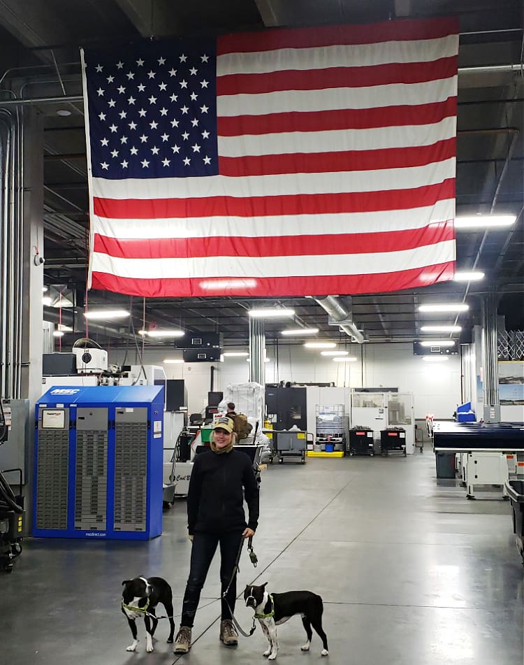 Sara Liberte with her two dogs standing under the huge American flag inside the SilencerCo plant.